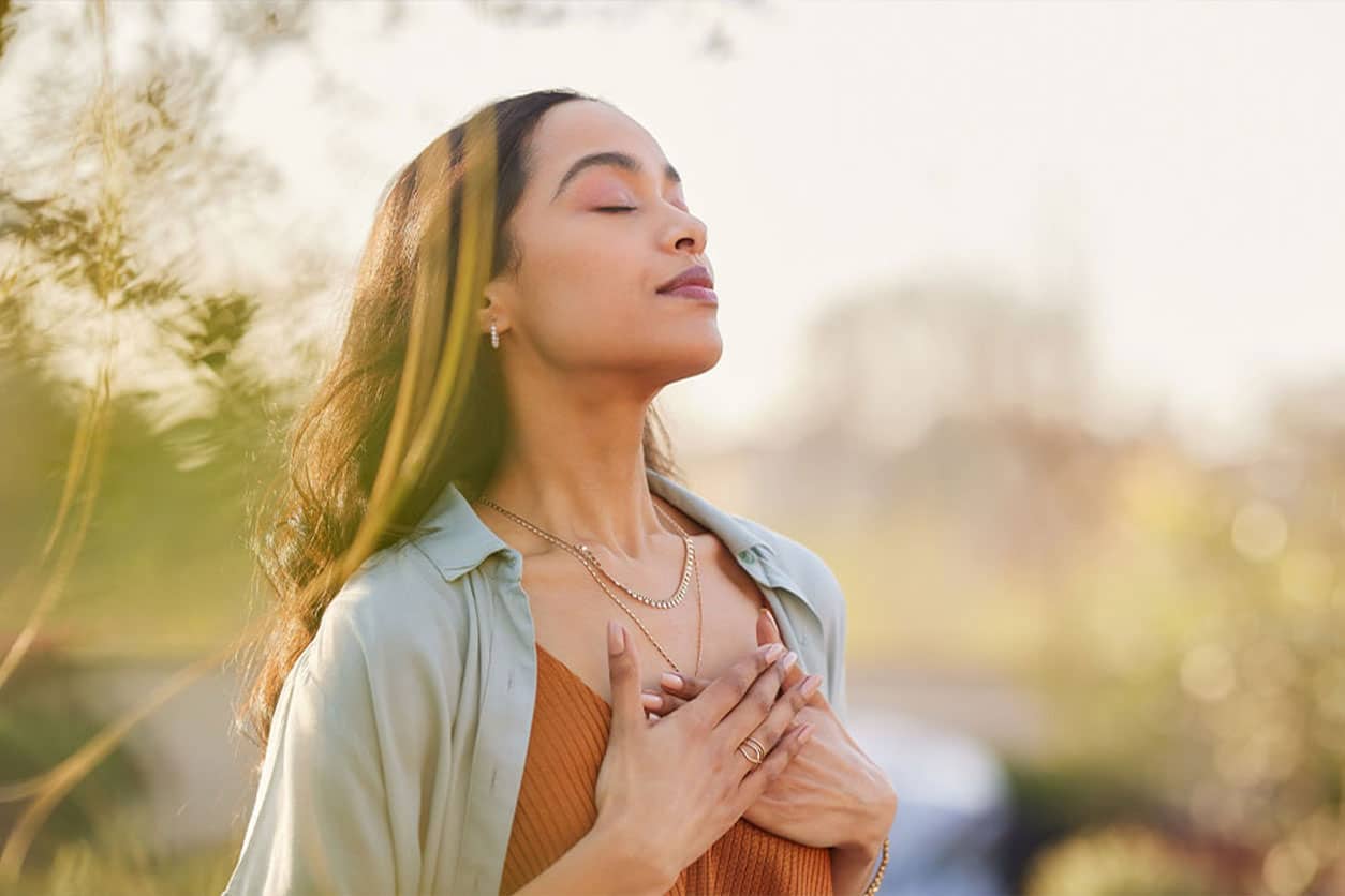 A woman practicing deep breathing for stress relief as she holds both of her hands over her heart and faces the sky.